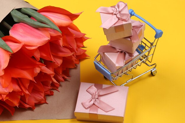 Buquê de tulipas e carrinho de compras com caixas de presente em fundo amarelo. Compras de férias. Dia das mães ou 8 de março, aniversário.