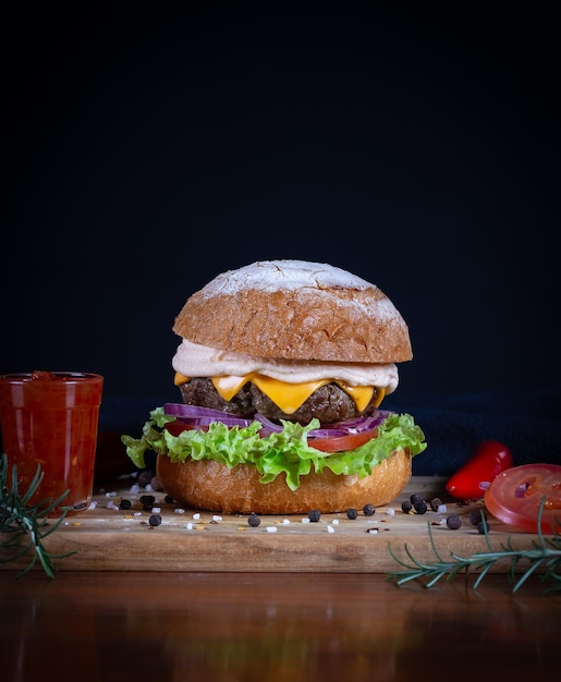 Foto burger de carne com alface, tomate, queijo cheddar e cebola servido em uma tábua de corte em uma mesa de madeira preta burger de carne artesanal feito à mão