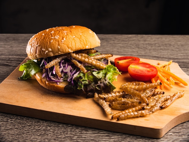 Burger mit gebratener Bambusraupe und Gemüsesalat auf Holzplatte.