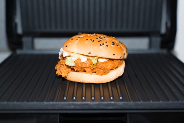 Burger mit Hühnchen paniert auf dem Elektrogrill, drinnen. Nahaufnahme, selektiver Fokus. Fast Food in der Küche zubereiten