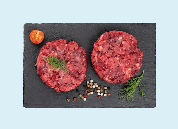 Burger Patties aus rohem Rinderhackfleisch auf schwarzem Schieferbrett mit Tomaten und Gewürzen