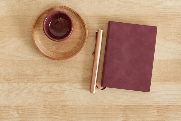 Burgunderrotes Notizbuch und Stift neben Tasse und Holzschale auf Holzschreibtisch