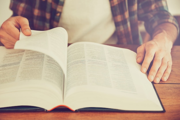 Buscando inspiración en un libro. Primer plano del hombre pasando la página del libro mientras está sentado en el escritorio de madera