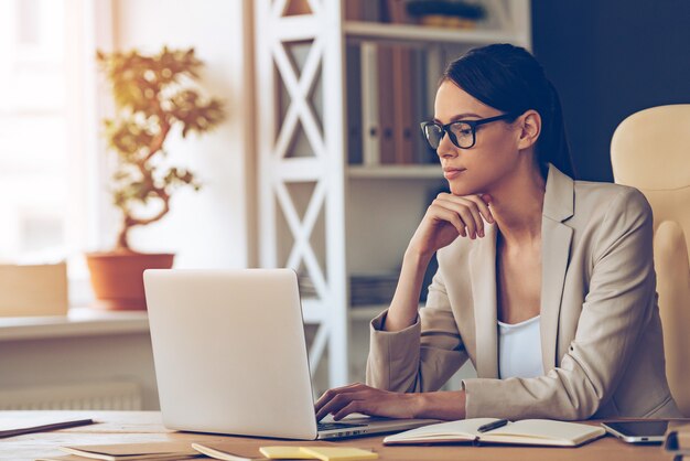 Buscando una nueva solución. Empresaria hermosa joven pensativa en gafas trabajando en la computadora portátil y manteniendo la mano en la barbilla mientras está sentada en su lugar de trabajo