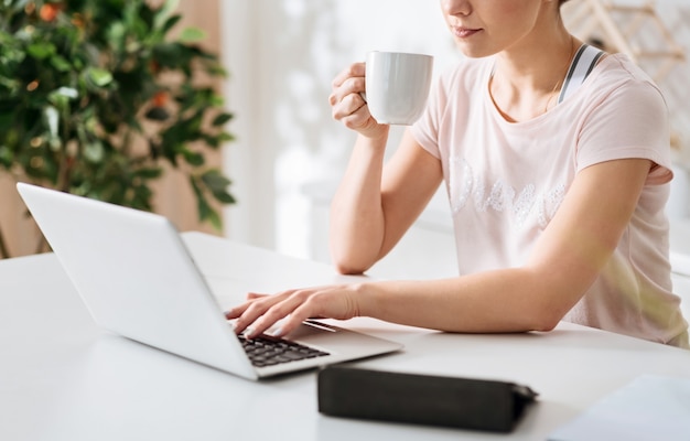 Buscando una solución. Bonita chica fácil pero enfocada usando su computadora navegando a través de algunas páginas web mientras toma un café por la mañana