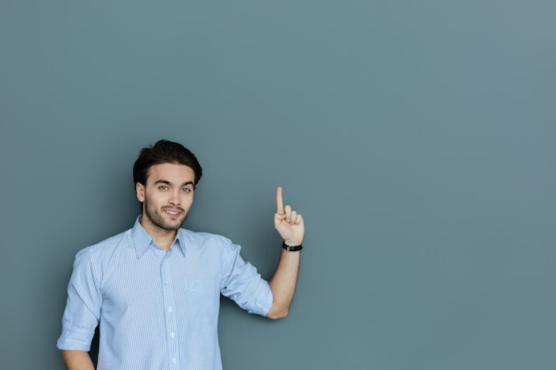 Buscar. Hombre creativo positivo alegre de pie contra el fondo gris y sonriéndole mientras apunta hacia arriba con su dedo