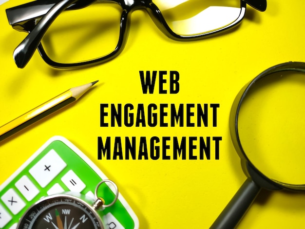 Business conceptText WEB ENGAGEMENT MANAGEMENT com calculadorabússolalápis lupa e óculos em fundo amarelo