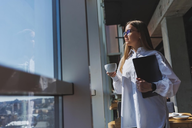 Business-Frau in einem weißen Hemd und Brille mit einem Laptop in der Hand im Büro am Fenster Moderne Geschäftsfrau in stilvoller Kleidung Freiberufliche Mädchen arbeiten außerhalb des Büros