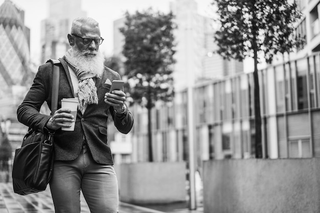 Business-Hipster-Senior-Mann benutzt Mobiltelefon, während er zur Arbeit geht, mit der Stadt im Hintergrund, Fokusgesicht, Schwarz-Weiß-Bearbeitung