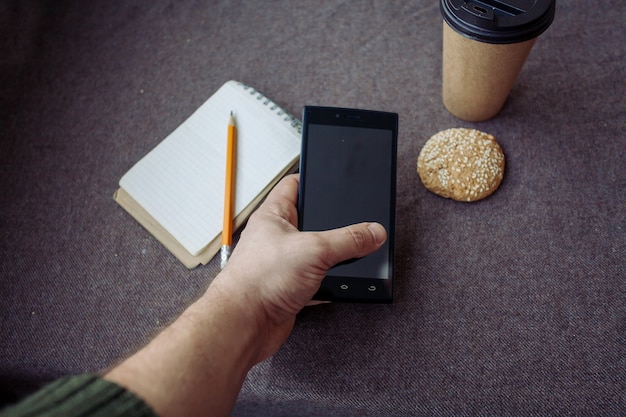 Business, Lifestyle, Essen, Menschen und Kaffeekonzept - Bleistift, Notizbuch und Papierkaffeetasse auf braunem Stoffhintergrund. Mann hält ein Telefon in der Hand