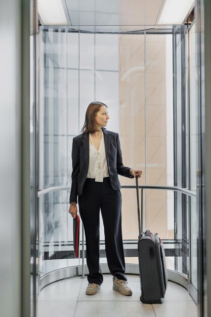 Business-Tourismus-Geschäftsfrau in einem formellen Anzug mit einem Smartphone in einem Business Center oder Flughafen mit einem Koffer geht auf Geschäftsreise