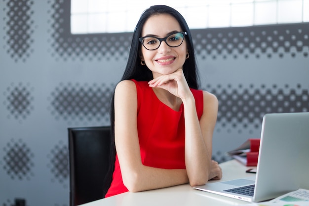 Businesslady atraente jovem de vestido vermelho e óculos sentar à mesa e trabalhar com laptop