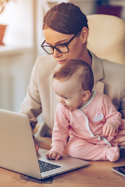 Businessplan mit Mama machen. Kleines Mädchen mit Laptop beim Sitzen auf dem Schreibtisch mit ihrer Mutter im Büro