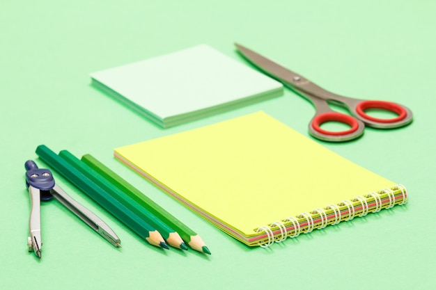 Bússola, lápis de cor, caderno, papel nota e tesoura sobre fundo verde.