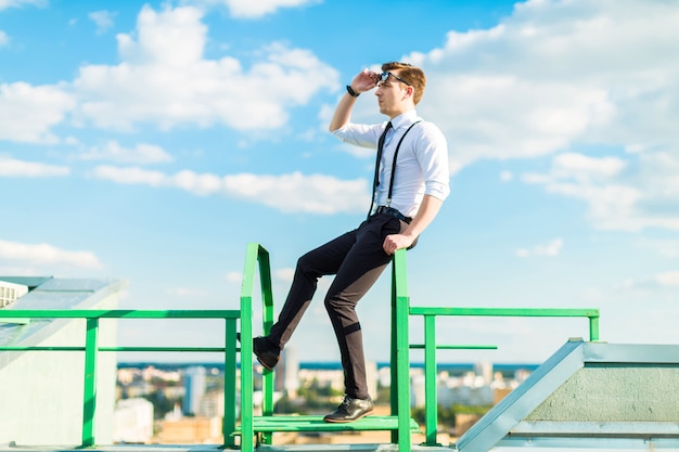 Busunessman jovem de camisa branca, gravata, aparelho e óculos de sol fica na escada do telhado