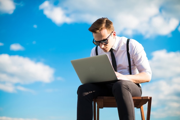 Busunessman sério atraente na camisa branca, gravata, aparelho e óculos de sol trabalha com o laptop no telhado