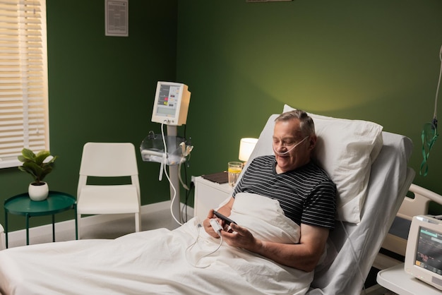 Un caballero anciano con un tubo de oxígeno y un monitor de oxigeno en el dedo habla por teléfono mientras se recupera en