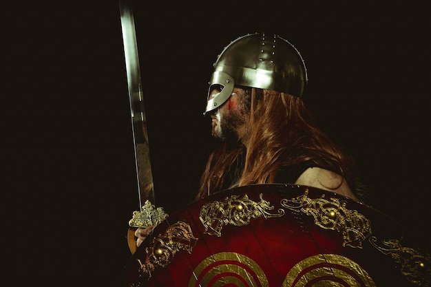 Caballero, guerrero vikingo con casco metálico y pieles de animales. guanteletes de cuero, escudo de madera rojo con adornos dorados y espada de acero