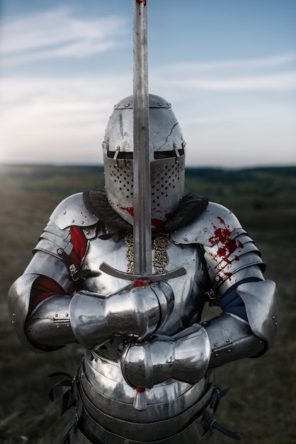 Foto caballero medieval con armadura y casco posa con espada, gran batalla. antiguo guerrero blindado con armadura posando en el campo