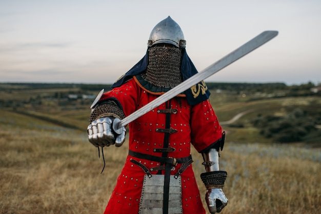 Foto caballero medieval en casco de metal tiene espada, gran batalla. antiguo guerrero blindado con armadura posando en el campo