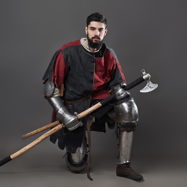 Foto caballero medieval sobre fondo gris. retrato de guerrero brutal cara sucia con armadura de cota de malla ropa roja y negra y hacha de batalla