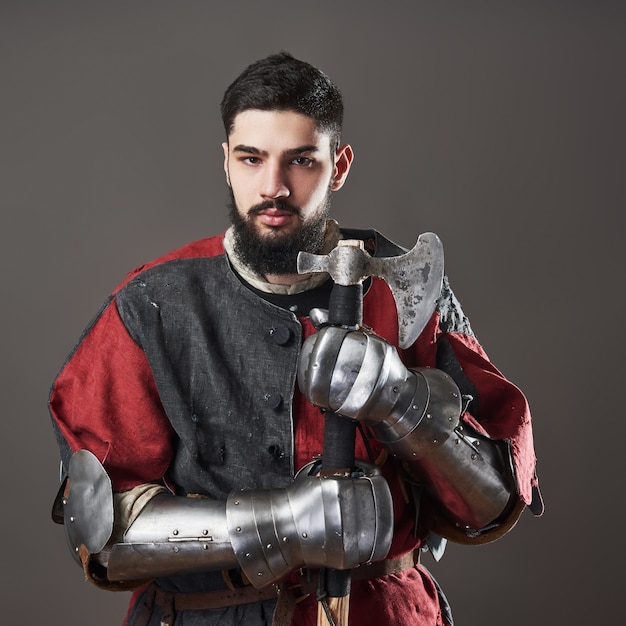 Foto caballero medieval sobre fondo gris. retrato de guerrero de cara brutal con armadura de cota de malla ropa roja y negra y hacha de batalla.