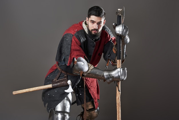 Foto caballero medieval sobre fondo gris. retrato de guerrero de cara brutal con armadura de cota de malla ropa roja y negra y hacha de batalla.