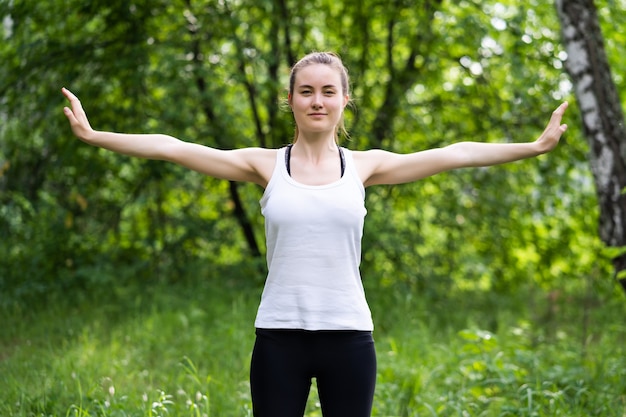 Cabe a jovem mulher bonita vestindo top branco e leggings esportivas pretas, malhando ao ar livre no parque num dia de verão.