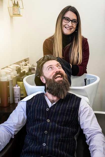 Cabeleireira secando o cabelo do cliente hipster depois de enxaguar no lavatório Homem hipster sorridente no salão de barbearia cortando barba e cabelo Conceito de barbearia