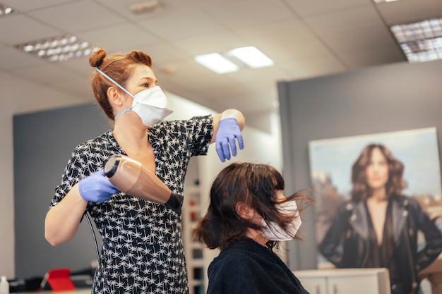 Cabeleireiro com medidas de segurança para a covid-19, nova normalidade, distância social. Secar o cabelo de uma cliente morena com uma máscara