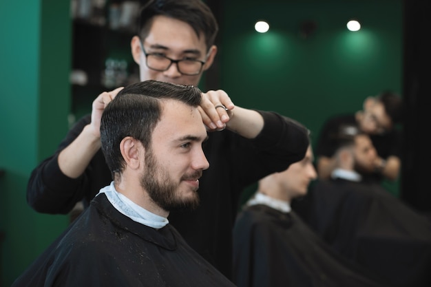 Cabeleireiro com um pente e uma tesoura corta um homem barbudo em uma poltrona em uma barbearia