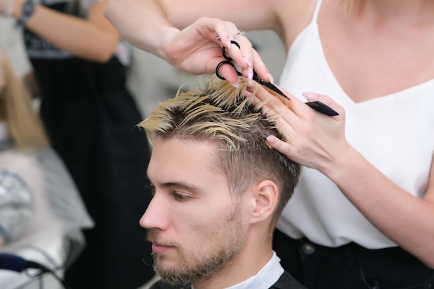 Cabeleireiro corta o cabelo de um jovem com cabelos tingidos de loiro com uma tesoura em um salão de beleza. cuidado profissional do cabelo.