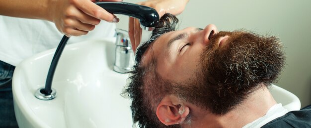 Cabeleireiro lavando o cabelo de seu lindo cliente. Limpeza de cabeça de barbearia. Homem bonito com barba na barbearia.