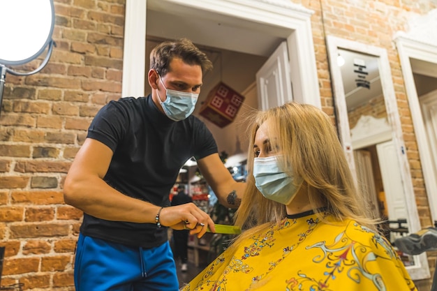 Cabeleireiro masculino com máscara penteando o cabelo loiro de suas clientes femininas no salão de cabeleireiro profissional