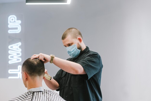 Cabeleireiro masculino em uma máscara protetora de gaze corta a máquina de cortar cabelo do cliente durante a quarentena Trabalho de um cabeleireiro durante a pandemia de coronavírus o barbeiro usa uma máscara médica