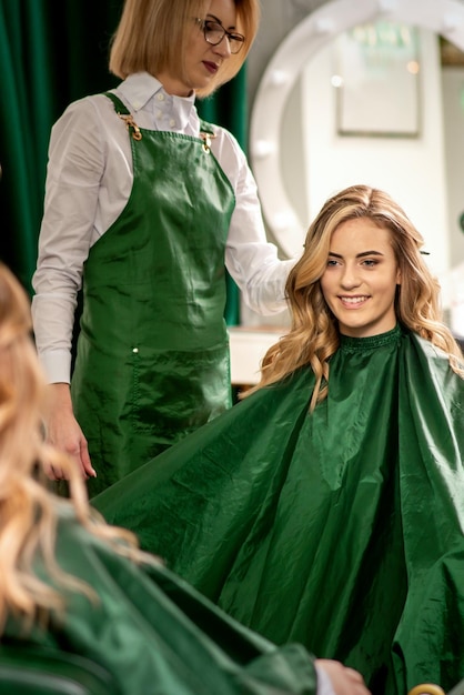 Cabeleireiro penteando o cabelo comprido de uma jovem mulher caucasiana olhando e sorrindo no espelho no salão de beleza