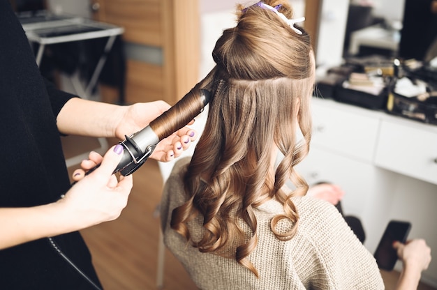 Cabeleireiro prepara mulher jovem e bonita para o evento, faz penteado cachos com um modelador de cachos para cliente no salão de beleza