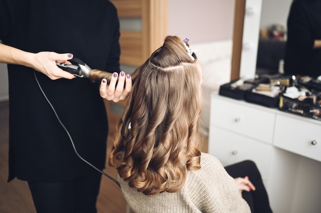 Cabeleireiro prepara mulher jovem e bonita para o evento, faz penteado cachos com um modelador de cachos para cliente no salão de beleza