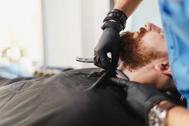 Cabeleireiro profissional atendendo cliente, barbeando uma barba espessa com navalha reta