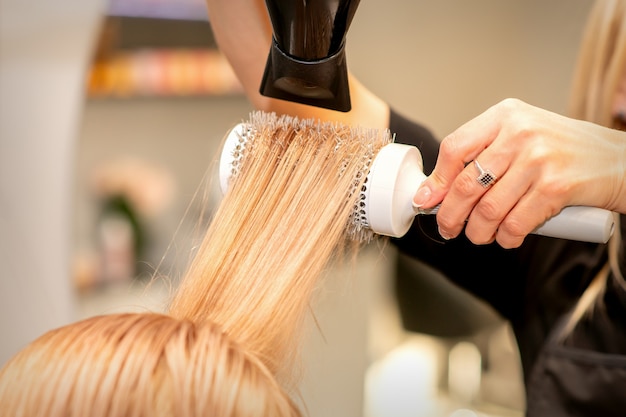 Cabeleireiro profissional seca o cabelo com secador de cabelo e escova de cabelo redonda em um salão de beleza.