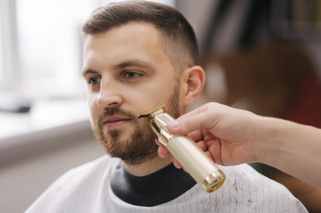 Cabeleireiro profissional usa um aparador de cabelo para franjar a barba de um homem bonito na barbearia