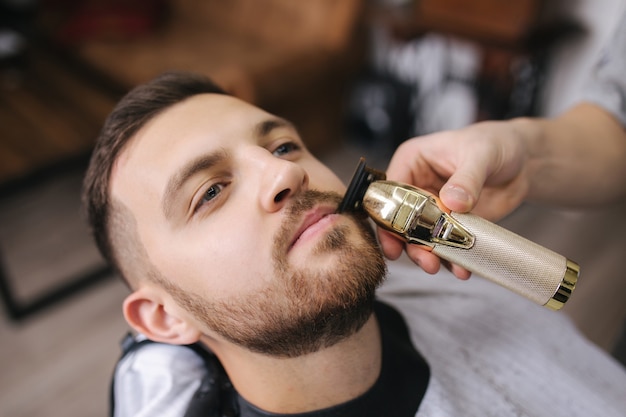 Cabeleireiro profissional usa um aparador de cabelo para franjar a barba de um homem bonito na barbearia.