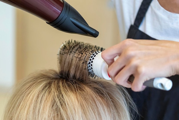 Cabeleireiro seca os cabelos molhados, faz a modelagem do cabelo em um secador de cabelo e penteia para uma cliente loira
