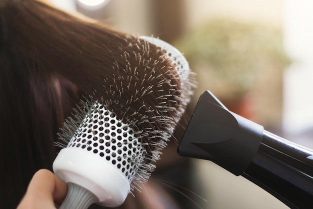 Cabeleireiro secando o cabelo de mulher com secador de cabelo e escova redonda no salão de beleza, close up