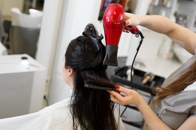 Cabeleireiro trabalha no penteado feminino no salão Secagem de cabelos castanhos compridos com secador de cabelo e escova redonda