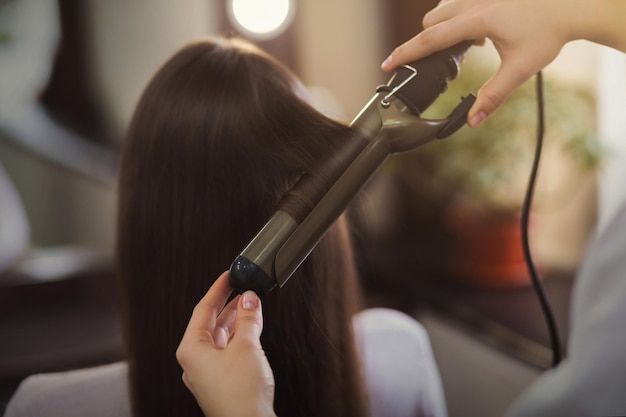 Cabeleireiro usando modelador de cabelo em longos cabelos castanhos em um salão de beleza