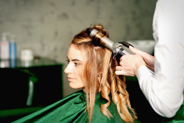 Cabeleireiro usando pinça de ondulação e cachos longos cabelos castanhos na jovem caucasiana em um salão de beleza