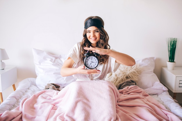 Foto cabello oscuro hermosa joven morena despierta en su cama. mujer agradable alegre que sostiene el reloj en manos y sonrisa. mira directamente a la cámara. máscara para dormir en la frente. modelo feliz positivo en el dormitorio.