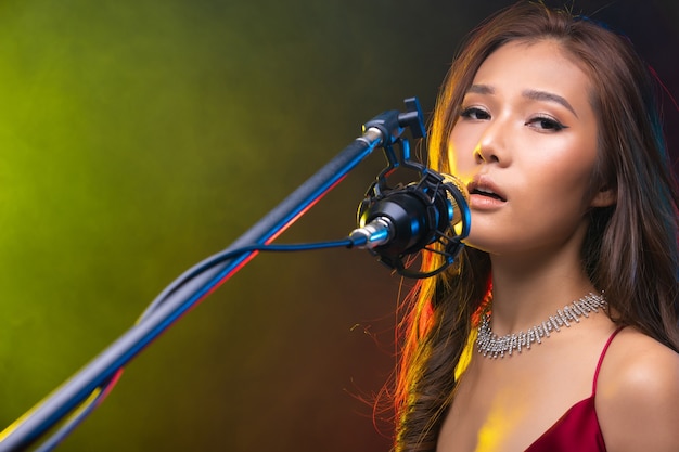 Cabelo castanho cacheado de mulher asiática usa vestido vermelho e canta uma música alto poder som sobre condensador de microfone pendurado, compositor. Absorvendo fumaça no estúdio, copie o espaço