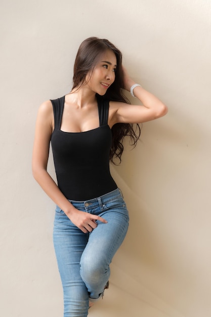 Cabelo castanho comprido de mulher bonita, sorrindo com uma camiseta preta e calça jeans em pé postando na parede de cor bege.